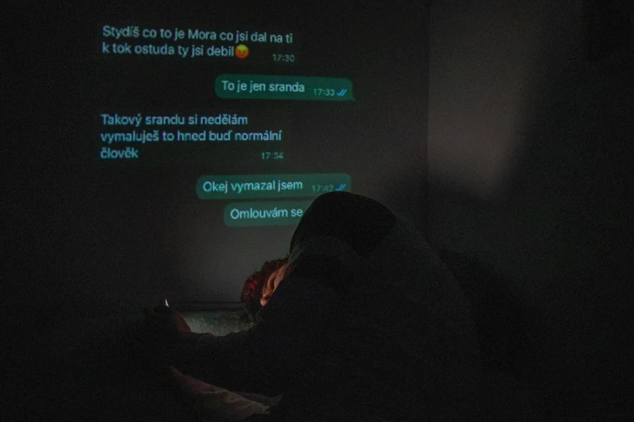 Autoportrét schoulený v posteli s projekcí zpráv na stěně.